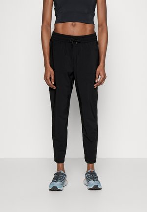 Person trägt schwarze Kordelhose, schwarzes kurzes Top und blaue Laufschuhe und steht vor einem einfarbigen Hintergrund.