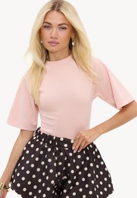Femme blonde portant un haut rose pâle à manches courtes et un short noir taille haute à pois blancs, posant avec la main sur la hanche.