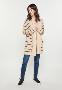 Béžový cardigan s černými pruhy, otevřený vpředu, dlouhé rukávy a kapsy. Spárován s modrými skinny džíny a černými teniskami.