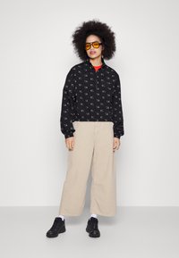 Jachetă neagră cu guler, cu un model alb, cămașă roșie dedesubt, pantaloni bej cu picioare largi, adidași negri și ochelari de soare oversized galbeni.