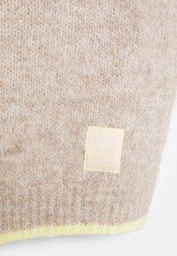Beige Strickmütze mit einer weichen Textur; verfügt über einen gelben Akzent entlang des Randes und ein Etikett mit der Aufschrift "Seit 1991".