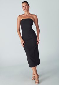 Melnā strapless midi kleita ar pieguļošu siluetu, gludu audumu, līdz ceļiem garu malu un vertikālo šuvju detaļām. Apvienota ar caurspīdīgām papēžkurpēm.