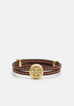 Brown leather bracelet with white stitching and a round gold-tone emblem featuring a symmetrical logo design at the center.
