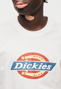 Λευκό φούτερ με πολύχρωμο στρογγυλό λογότυπο που περιλαμβάνει τη λέξη "Dickies" σε μπλε χρώμα, πάνω σε κόκκινο φόντο.