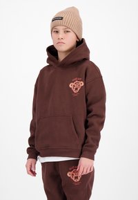 Jongen draagt een bruine hoodie en bijpassende joggingbroek met cartoonbeer-logo en een beige geribbelde gebreide muts, staand tegen een witte achtergrond.