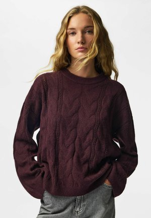 Jeune femme aux longs cheveux blonds portant un pull torsadé violet foncé et un pantalon gris, mains dans les poches, fond neutre.