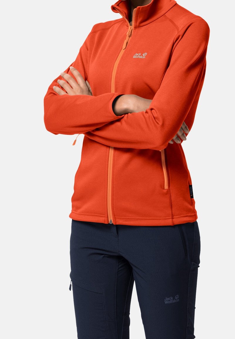 Osoba w pomarańczowej kurtce zip-up z założonymi rękami i ciemnoniebieskich spodniach, obie części odzieży z logotypem Jack Wolfskin.