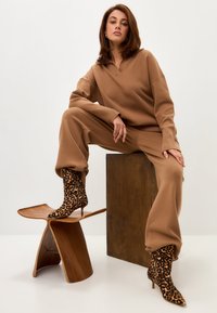 Pulover maro �și pantaloni dintr-un material moale, având un guler polo. Botine cu imprimeu animal și tocuri stiletto. Taburet din lemn și fundal din bloc.