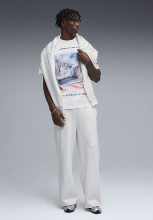 Mann trägt weißes Grafik-T-Shirt mit dem Schriftzug "RISK IT ALL", weiße Jogginghose und einen Pullover über den Schultern drapiert, steht vor grauem Hintergrund.