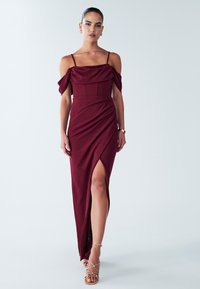 Burgundská večerní šaty s širokými rameny, přiléhavým korzetem, draped sukní a rozparkem po stehna, vyrobené z hladkého materiálu. Doplněné o páskové podpatky.