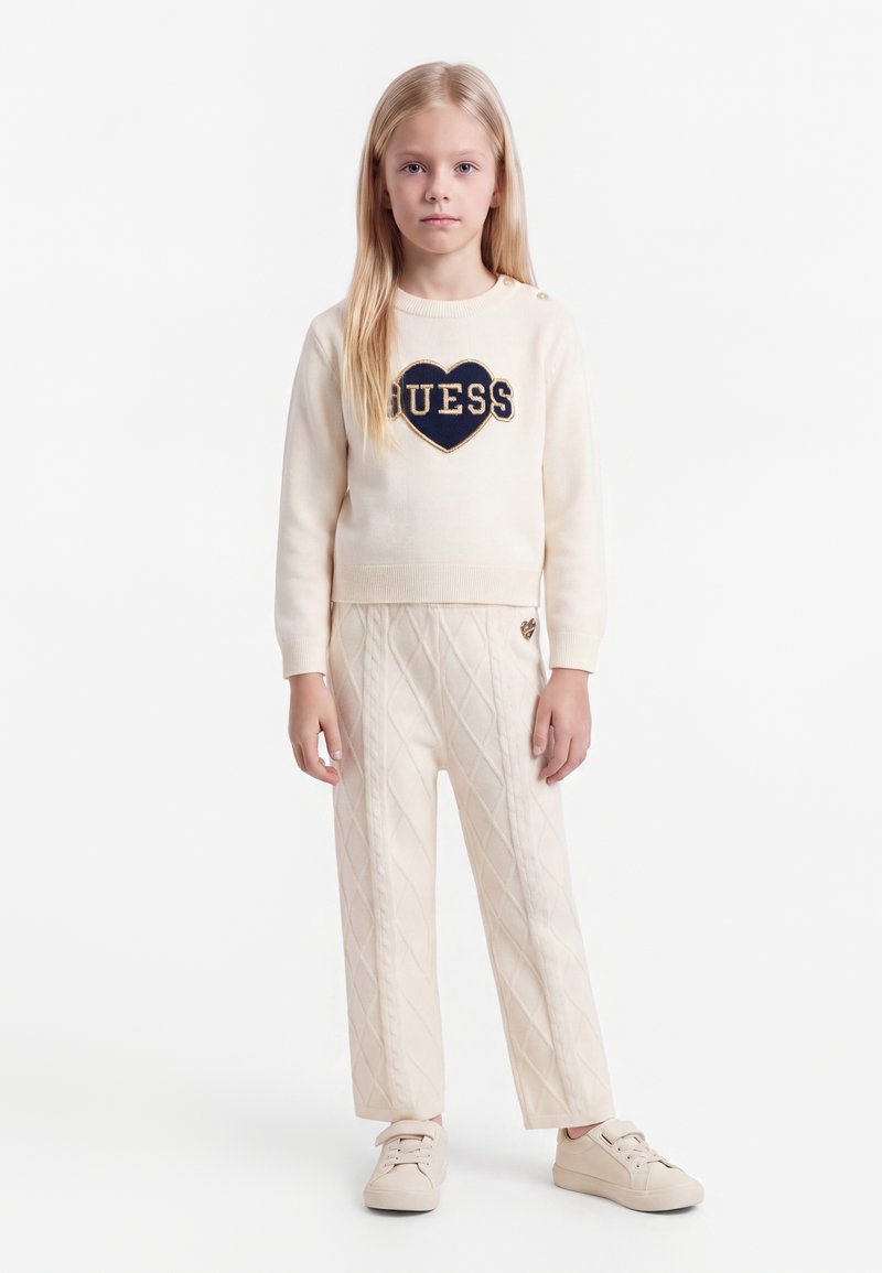 Jeune fille aux longs cheveux blonds portant un pull crème avec un cœur bleu marine et le texte "GUESS", pantalon matelassé assorti et baskets claires, debout.