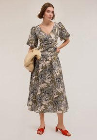 Abito a stampa floreale verde oliva con maniche a sbuffo, scollo a V e vita arricciata. La modella tiene una borsa beige e indossa sandali rossi con tacco.