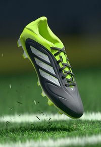 Schwarzer und neongrüner Fußballschuh mit drei weißen Streifen, strukturiertem Obermaterial und genopptem Sohlen, präsentiert über Gras mit Erdpartikeln.