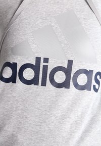 Sudadera gris de algodón con un gran logo de Adidas en gris claro liso y texto en azul marino oscuro. Diseño sencillo sin patrones adicionales.
