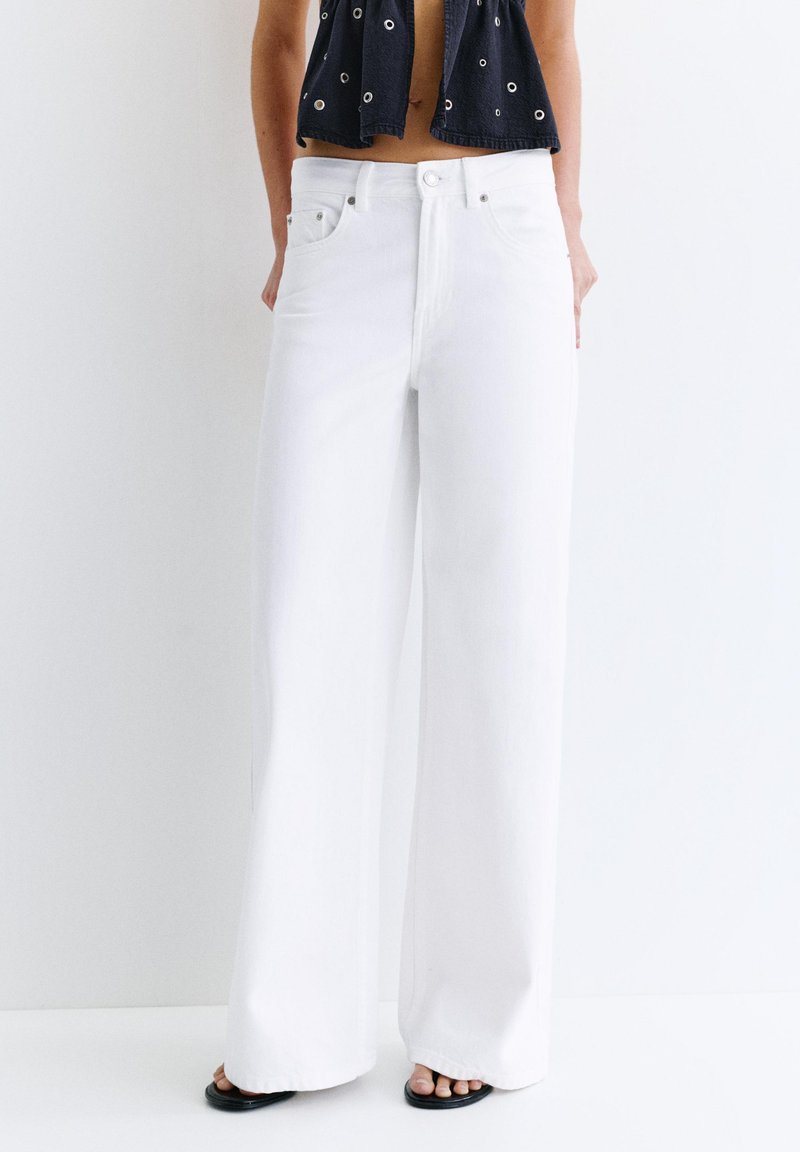 Personne portant un jean blanc à jambes larges, des sandales noires ouvertes aux orteils et un top court noir avec des détails œillets, debout devant un fond uni.