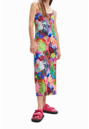 Vrouw draagt een mouwloze, getailleerde midi-jurk met een helder meerkleurige bloemenprint en roze sandalen met open tenen en zwarte zolen.