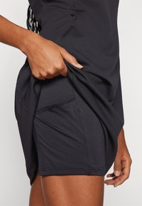 Μαύρο αθλητικό φόρεμα με πλαϊνή τσέπη και υφασμάτινη υφή, με χέρι που κρατά το ύφασμα κοντά στη μέση, αναδεικνύοντας την ευκαμψία.