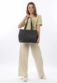 Sort tekstureret tote-bag med lange håndtag, der har et minimalistisk design. Tasken holdes af en model iført lysfarvede tøj imod en hvid baggrund.