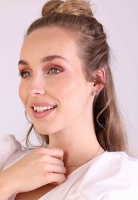 Modèle portant un simple collier en argent et des boucles d'oreilles clous. Cheveux coiffés en chignon, et le maquillage présente des tons chauds avec une légère brillance.