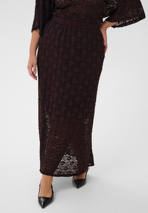 Femme portant une jupe midi en dentelle violette foncée avec un motif floral et des talons noirs pointus, debout avec une main sur la hanche.
