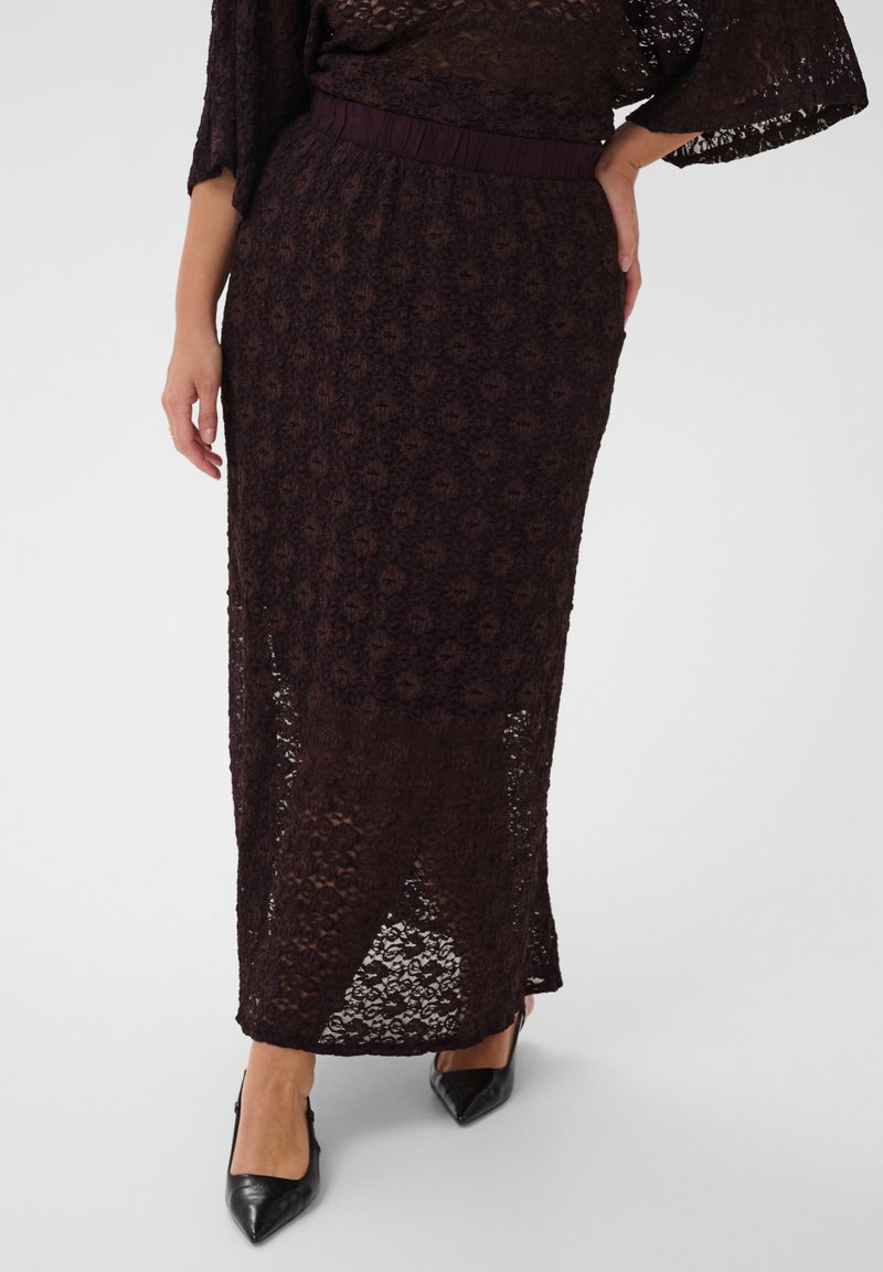 Femme portant une jupe midi en dentelle violette foncée avec un motif floral et des talons noirs pointus, debout avec une main sur la hanche.