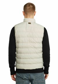Gilet imbottito di un beige chiaro, con colletto rialzato, texture a motivo trapuntato orizzontale e un'etichetta sul retro. Indossato sopra un top a maniche lunghe nero.