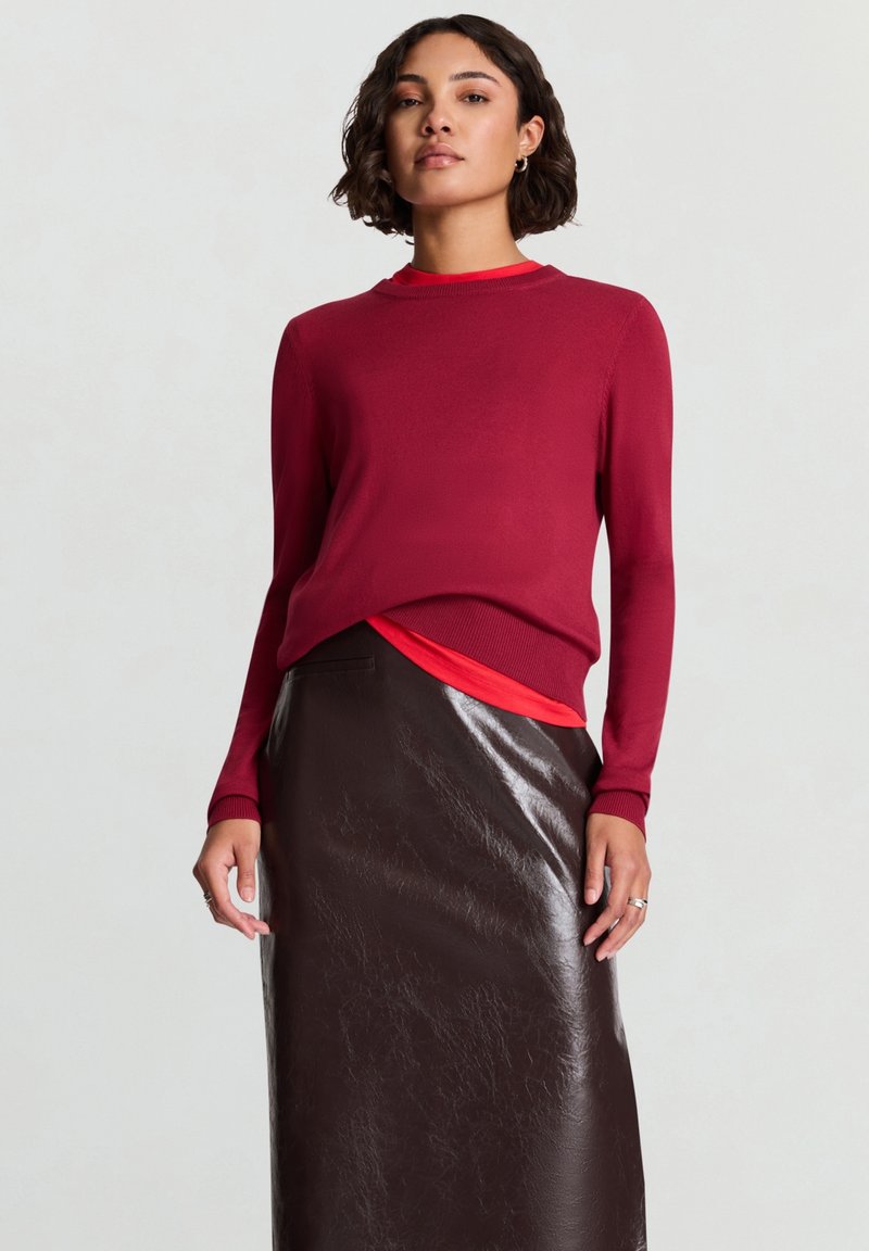 Bordeauxrotes langärmliges Oberteil trifft auf eine rote Akzentschicht; kombiniert mit einem hoch taillierten braunen Rock aus Kunstleder. Der Kontrast der Texturen zwischen den Stoffen wird hervorgehoben.
