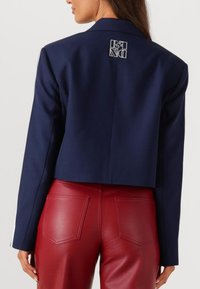 Marineblauwe cropped blazer met een gladde textuur en scherpe revers. Heeft een bedrukte ontwerp op de achterkant, gecombineerd met rode leren broek.