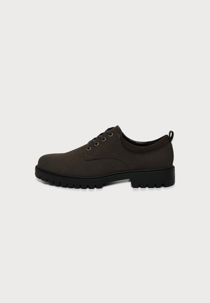 Chaussure en cuir marron foncé à lacets avec semelle épaisse en caoutchouc noir et languette au talon sur fond gris uni.