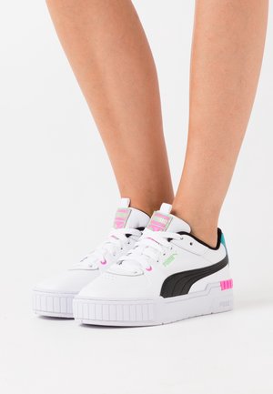 Baskets blanches avec des accents noirs et une semelle blanche épaisse. Les couleurs présentes incluent le rose et le sarcelle sur la languette et l'onglet du talon.