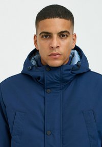Navy blue jacket with a hood, featuring a smooth texture, zip and button closures, and a grey interior lining. Structured design with pockets.