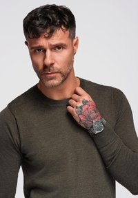 Maglione verde oliva con scollatura rotonda, accompagnato da un orologio d'argento e un tatuaggio di una rosa rossa sul polso.