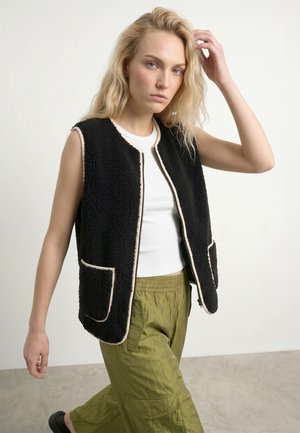 Donna bionda che indossa un gilet nero senza maniche con zip e bordo beige, una canottiera bianca senza maniche e pantaloni larghi verde oliva, si sistema i capelli con una mano.