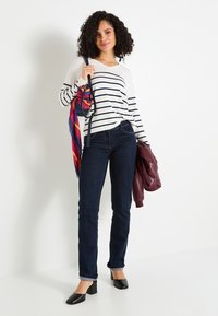 Mujer con pelo rizado que lleva un suéter a rayas blancas y negras y jeans oscuros, sosteniendo una chaqueta burdeos y un bolso con una bufanda colorida.