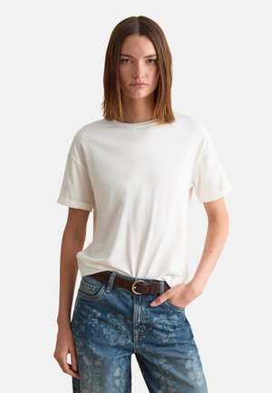 Vrouw met een eenvoudige witte T-shirt met korte mouwen, ingestopt in een hoge taille blauwe spijkerbroek met bloemenprint, staand met één hand in de zak.