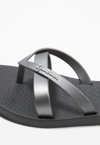 Graue Flip-Flops mit einer strukturierten schwarzen Sohle, Kreuzriemen und metallicfarbenem Markenlogo auf dem Riemen. Einfaches Design mit minimalen Details.