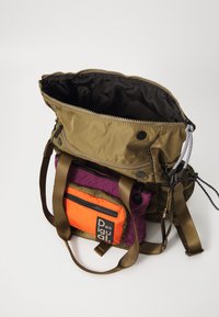 Mochila multicolor con un exterior marrón, acentos en púrpura y naranja, cierre de cremallera, múltiples correas y detalles de textura distintivos.