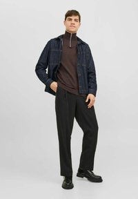 Veste en denim avec poches avant, pull-over marron, pantalon noir et bottines noires à lacets. Le modèle se tient devant un fond gris clair.