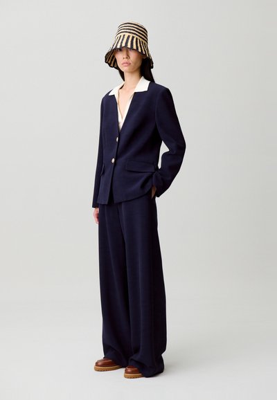 Abito su misura blu navy con colletto crema e bottoni dorati, abbinato a pantaloni a gamba larga e cappello tipo secchiello a righe intrecciato. Scarpe marroni.