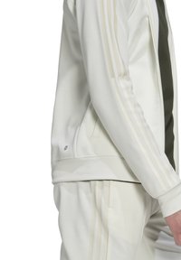 Biała sportowa kurtka z delikatnymi paskami na rękawach, z zapięciem na zamek i ściągaczami, w połączeniu z dopasowanymi dresami.