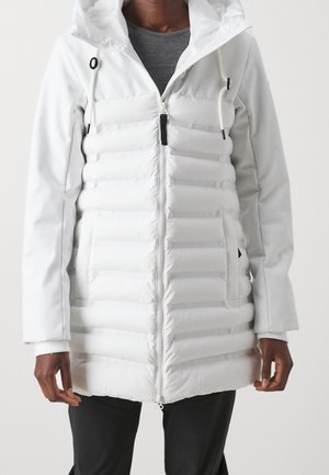 Personne portant une veste matelassée blanche avec capuche, fermeture éclair devant, cordons et poches latérales sur une chemise grise et un pantalon noir.