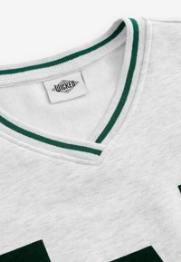 Biała bluza z dekoltem w serek ozdobiona zielonymi paskami, teksturowanym materiałem oraz metką z logo w okolicy kołnierza.