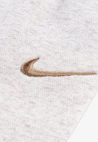 Hellbeiger Stoff mit einer strukturierten, gewebten Optik, der in einer unteren Ecke ein goldenes, besticktes Nike-Swoosh-Logo aufweist.