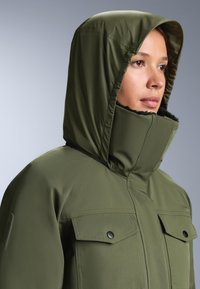 Olivno zelena jakna s kapuco in visokim ovratnikom, ki ima dve veliki sprednji žepni s pritrdilnimi gumbi ter gladko, vodoodporno teksturo tkanine.