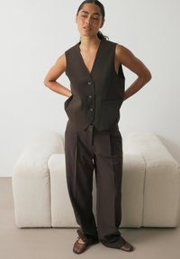 Rjav brezrokavnik z globokim V-izrezom, dvema sprednjima žepoma in tremi gumbi, v kombinaciji z ohlapnimi rjavimi hlačami.