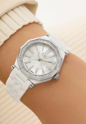 Reloj octagonal blanco con esfera de madreperla, acentos plateados y adornos de piedras preciosas transparentes, presentado en una muñeca.