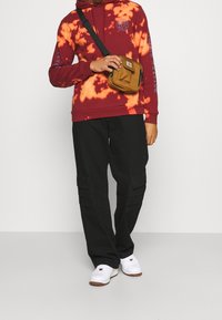 Felpa tie-dye rossa e arancione con logo frontale, abbinata a pantaloni cargo neri con tasche laterali e sneakers bianche. Borsa a tracolla marrone.