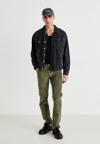Young man stands wearing a black denim jacket, black tank top, olive green pants, black shoes, and gray cap with white text on white background.