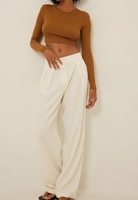 Kvinde iført en brun langærmet cropped bluse og højtaljede vidde bukser i hvid, stående foran en beige baggrund.