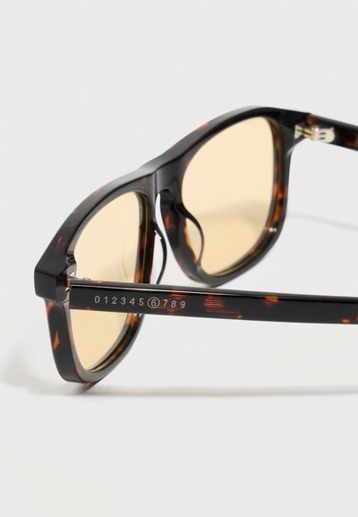 Γυαλιά ηλίου με κέλυφος χελώνας και φακούς με κίτρινη απόχρωση, με αριθμητική σήμανση 0123456789 στο μπράτσο του σκελετού, σε λευκό φόντο.